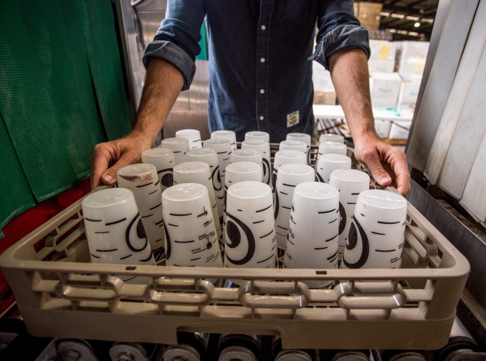 Sustainable Strategies | A person placing a tray full of dirty plastic cups into an industrial dishwasher.